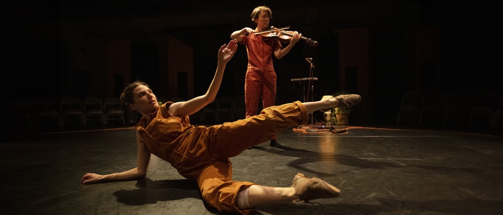 Two female performers appear in a black box space in burnt orange jumpsuits. 