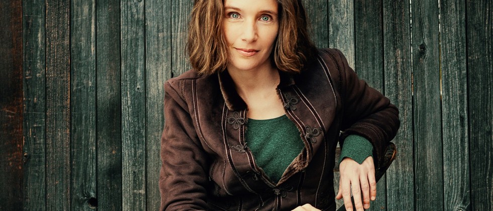 A green-eyed, brown-haired white woman. She smiles slightly and wears jeans, a brown suede jacket, and a green-blue wool sweater.