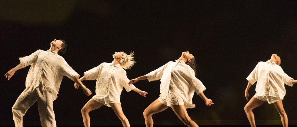 A dance company, men and women in ivory linen rompers, stepping forward, gesturing backward, and looking up in unison