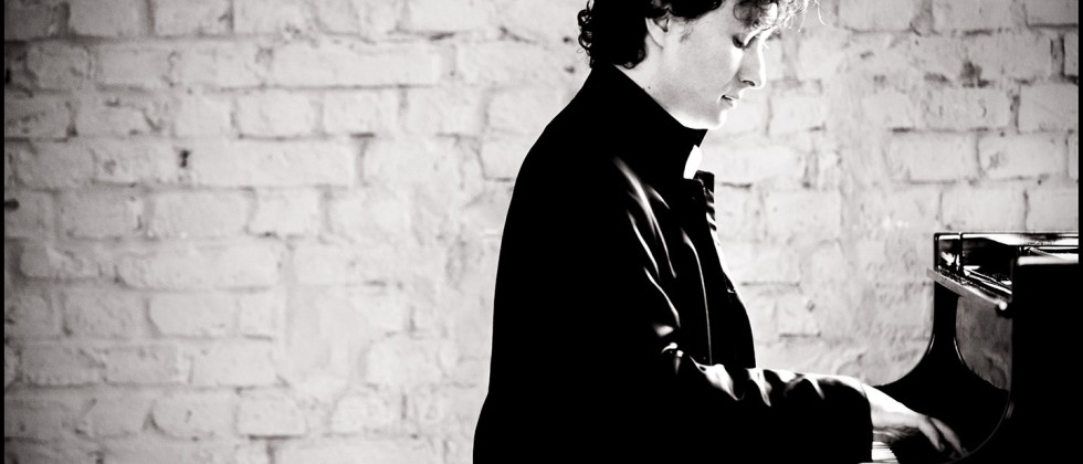 a straight-backed young white man with wavy dark hair plays a piano in front of a painted brick wall