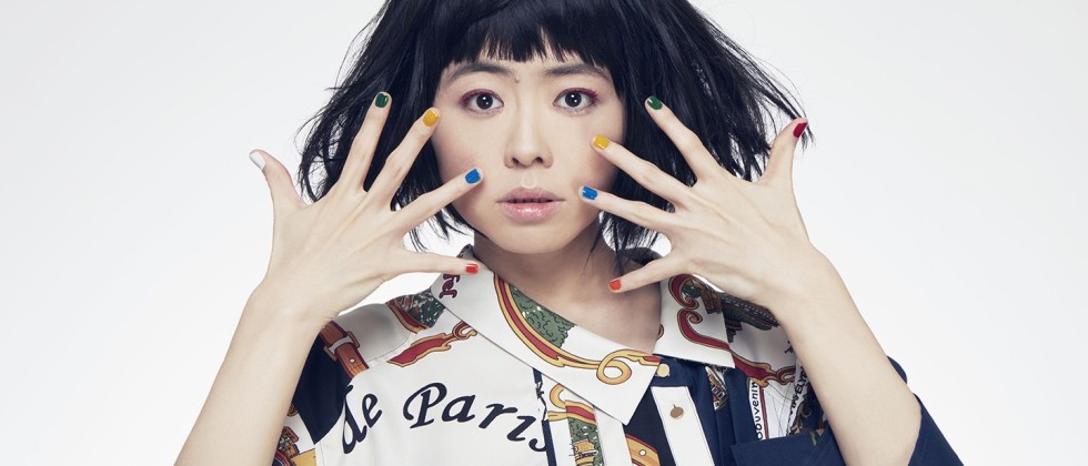 Hiromi, an Asian woman with dark hair wearing a patterned shirt holding her hands in front of her face in front of a white background. 