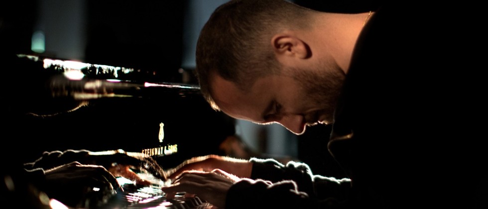 Igor Levit, a white man in a black top plays the piano with his head bowed. 