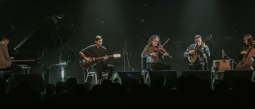 Five musicians are lined up in front of a large crowd playing a variety of instruments with two spotlights shining down on them. 