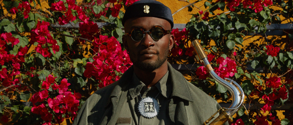 Isaiah Collier, a black man in his 20s, wears an olive green jumpsuit with an olive overcoat draped like a cloak, sunglasses, and a short lustrous cap