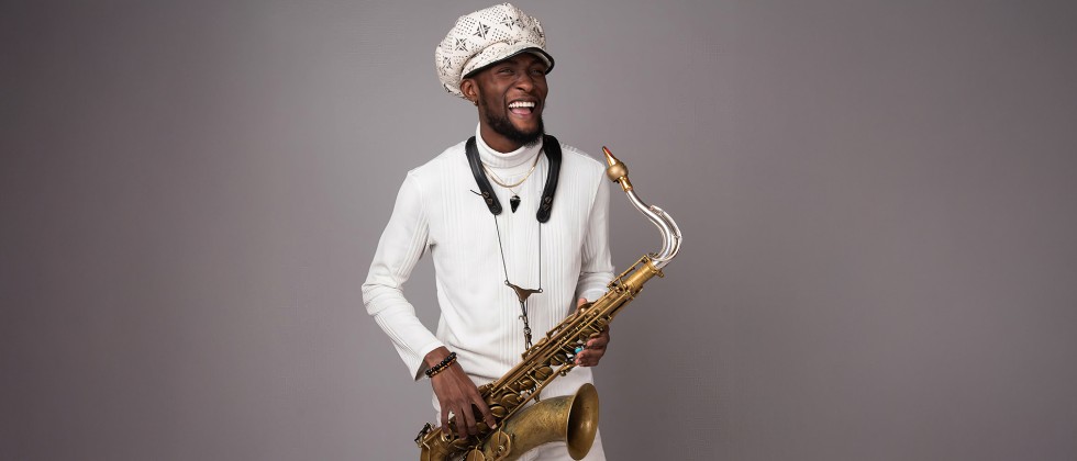 A young black man wearing an oversized paperboy cap stands holding a tenor saxophone with a vintage finish. 