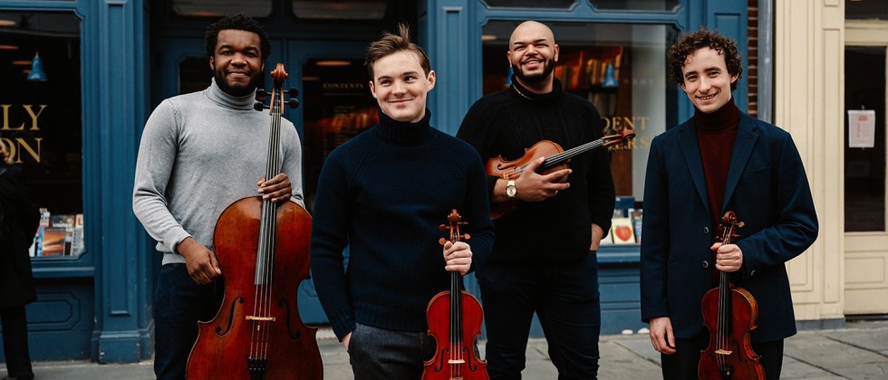 four young men smile impishly as they hold their string instruments in front of a bookstore with a bright blue facade.