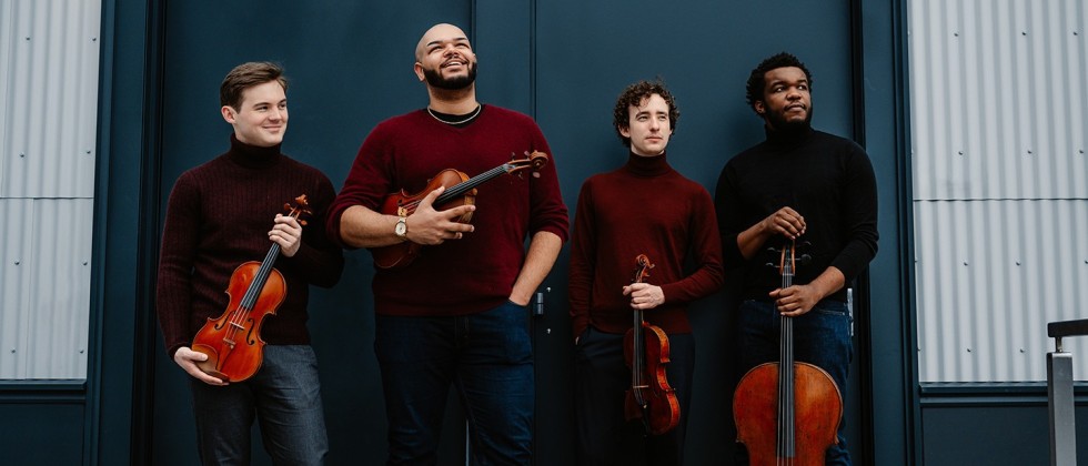 four young men smile impishly as they hold their string instruments in front of a bookstore with a bright blue facade.