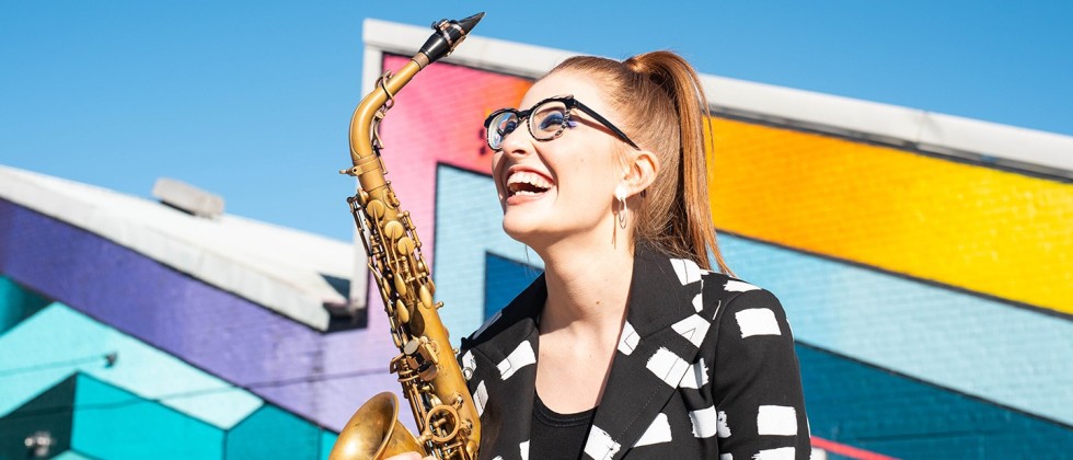 A young white woman with glasses and her red hair in a sleek ponytail, smiles broadly as she holds her tenor saxophone in front of a colorful geometric wall.