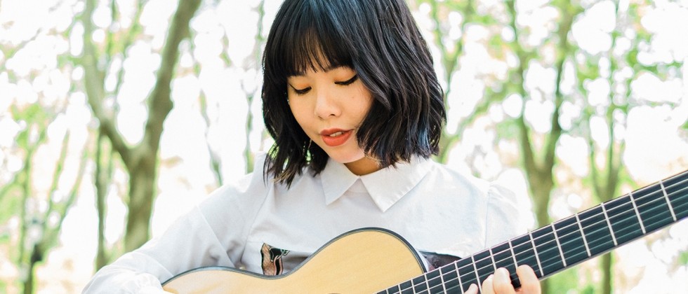 a young East Asian woman with blue-tinted wavy black hair looks down at her guitar as she plays outdoors among the trees