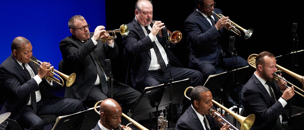Four members of a jazz band are shown playing brass instruments in suits with polka dot ties on a stage with a blue background. 