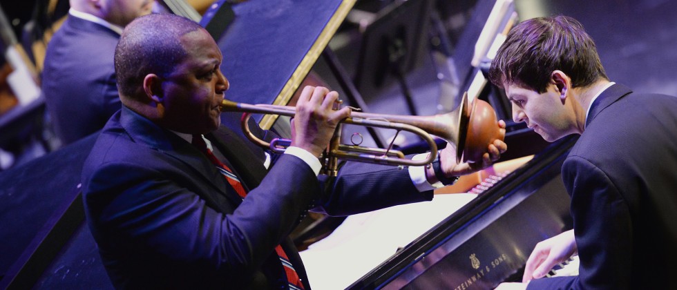 A grand piano with a white man playing fills the background, while a Black man plays trumpet with a mute in the foreground.  