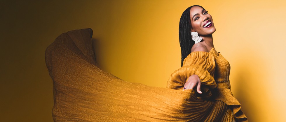 A black woman flourishes a pleated yellow dress while smiling widely in front of a shadowy yellow background. 