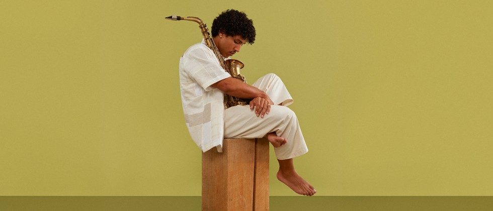 A Puerto Rican man in a white flowing outfit sits on a small wooden box, legs pulled up towards his body, cradling a saxophone. 