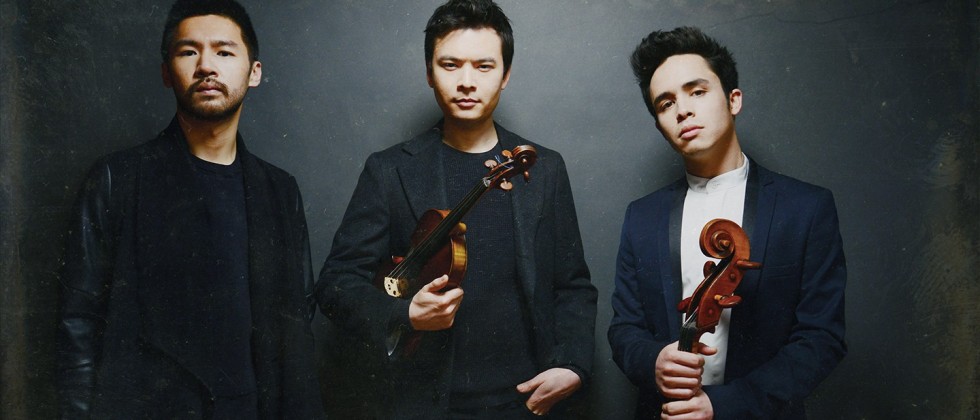 three young men stand in front of a gray portrait background, one holding a violin and one a cello.