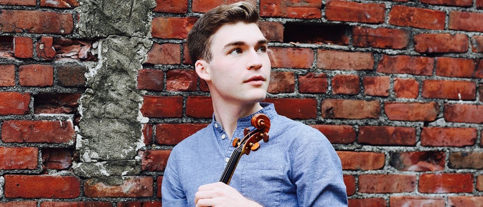 Alexi Kenney a white man in a blue shirt stands in front of a brick wall holding a violin. 