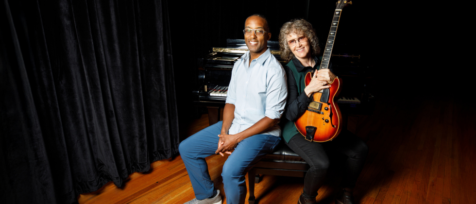 A black man in jeans and a casual buttondown leans against a white woman holding a hollowbody electric guitar. They sit on a bench in front of a grand piano