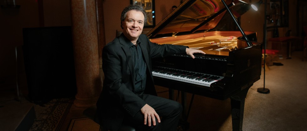 A white man in a black suit with swept back gray hair sits at a grand piano with one arm leaning on the rim over the keys. 