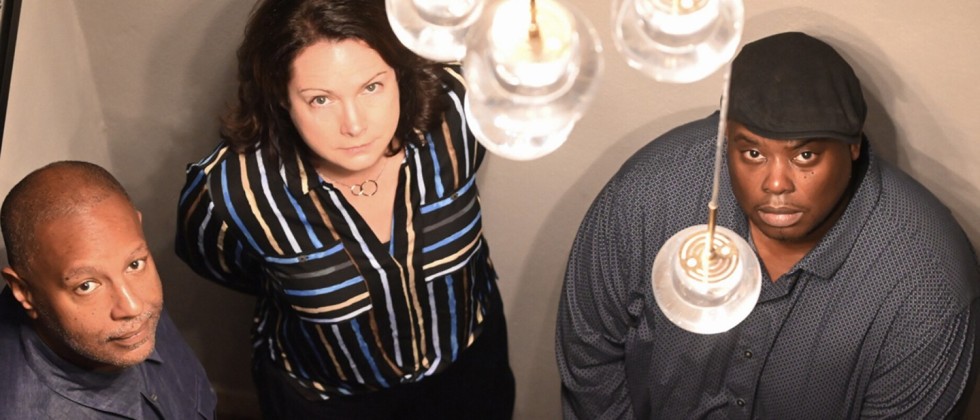 A white woman stands between two black men; the trio looks up with stoic expressions towards a lighting fixture with clear globes. 