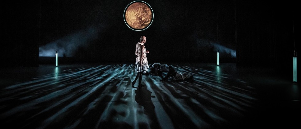 a male dancer stands alone on a dark stage, turned away from us toward two prone dancers and a moon-like illuminated circle