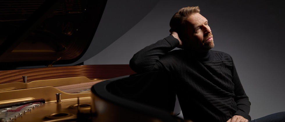 A white man in a black sweater sits next to a grand piano, leaning one arm against the curve of the piano.