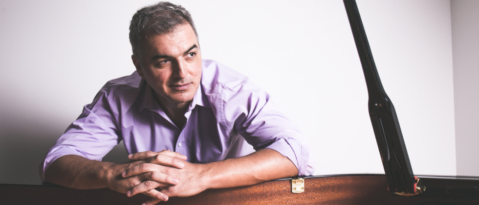 a latino man in a light purple shirt leans over the edge of a grand piano, his hands clasped