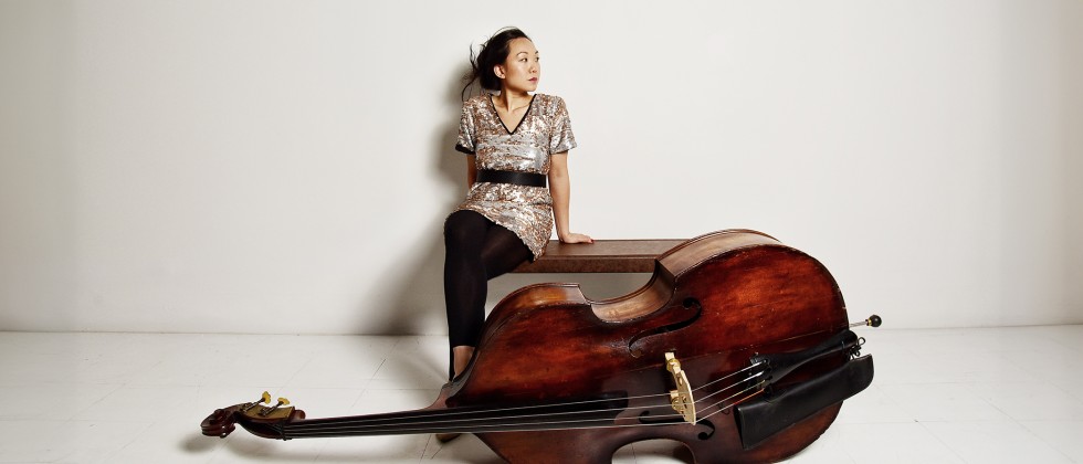 A Malaysian Australian woman sits on a bench with her legs crossed, on the floor leans her bass, it’s reddish wood. 