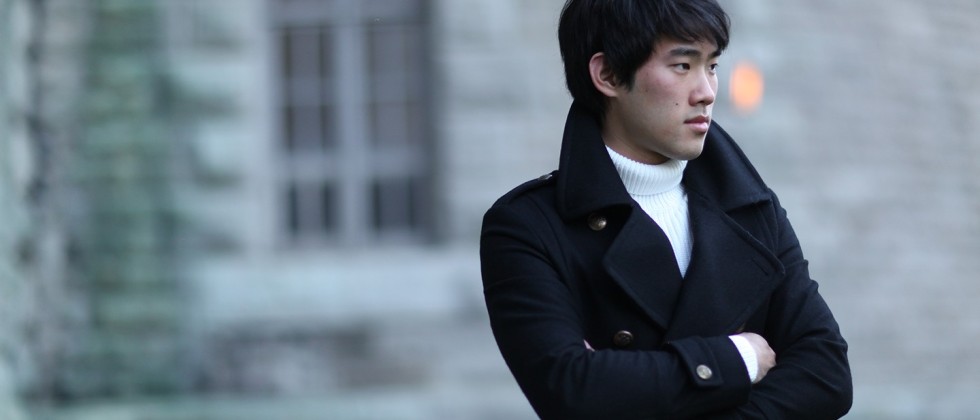 a young east asian man crosses his arms in front as he stands outdoors in front of a stone building. He wears a white turtleneck sweater and a pea coat.