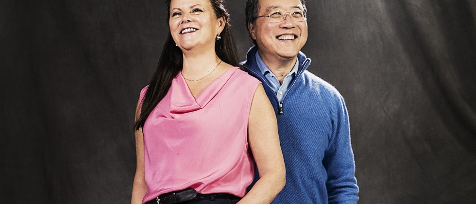 a white woman with long brown hair and a pink top and a gray-haired asian man with glasses and a quarter-zip blue pullover stand with their hands in their pockets, smiling happily. They look like they just shared an inside joke.