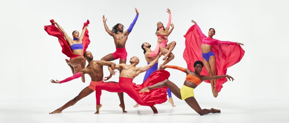 6 dancers gesture up and out, dressed in bright red and blue flowing material. 