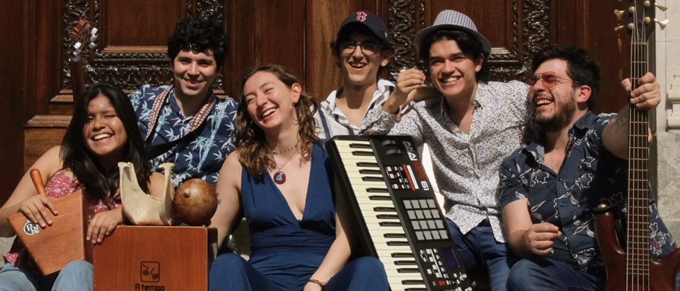 A mixed group of 6 joyful people sit and holding an electric guitar, keyboard, and percussion instruments on the steps outside ornate doors. 