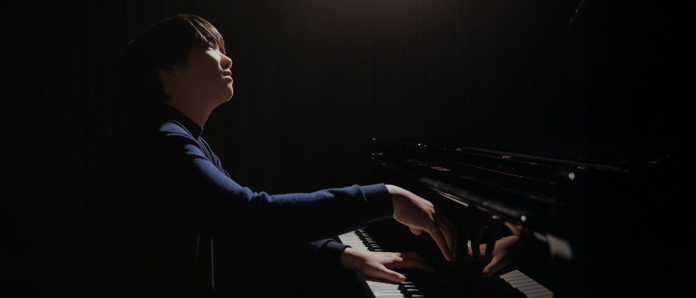 A young, Japanese man sits playing the piano as a spotlight shines from above. 