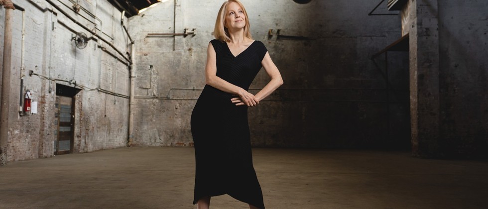 a blonde white woman in a black dress stands with a hand on her hip in an industrial building