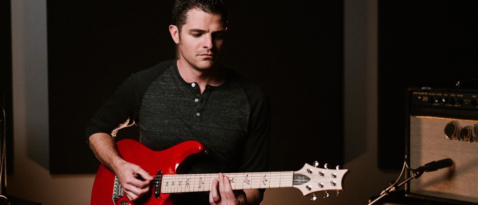 a white man looks down as he plays his red electric guitar in a music studio. His dark hair is slicked back and he has sideburns.