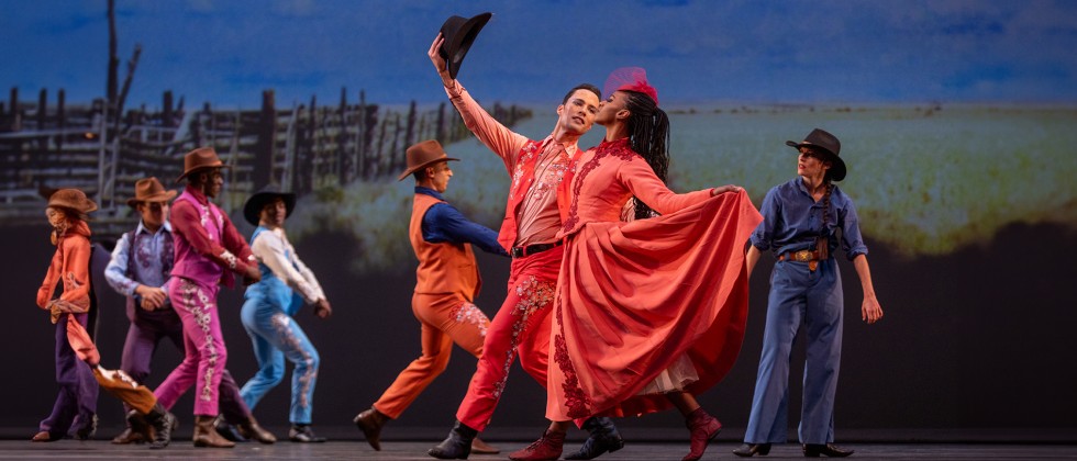 Before a western backdrop, a white male and Black female dancer perform in elaborate coral costumes with old western design.