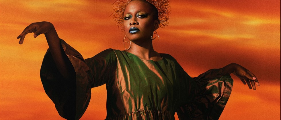 Crowned with a halo of tangled wire, a black woman in a metallic copper bell-sleeved dress floats her arms away from her sides. Her expression is enigmatic but wary, her makeup metallic and theatrical