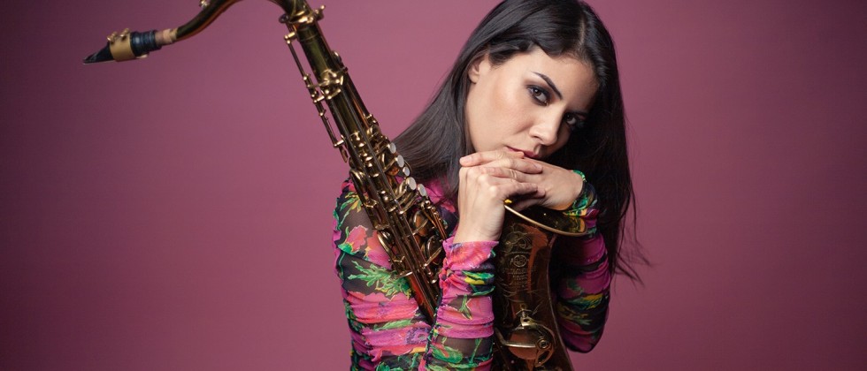 Melissa Aldana, a woman with brown hair sits holding a saxophone with her hands at her chin in front of a pink background.