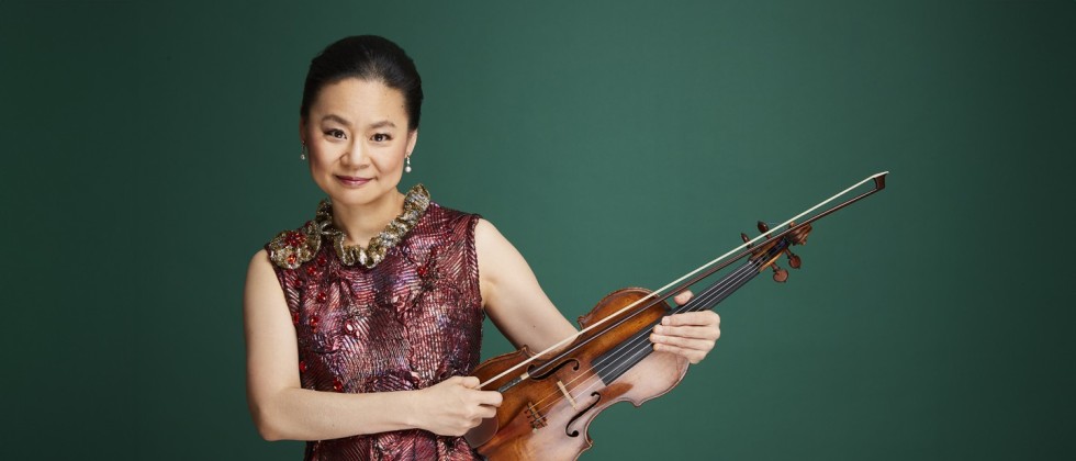 A Japanese woman stands her hair is pulled back wearing a textured red and gold gown, posed with her violin resting on her hip. 