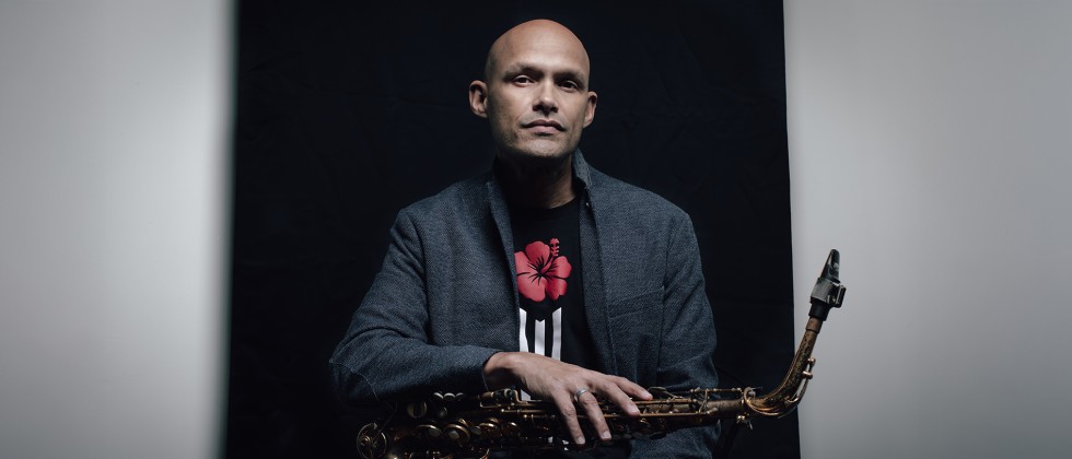 A Puerto Rican man in a gray jacket sits with a saxophone across his lap in front of a black background. 