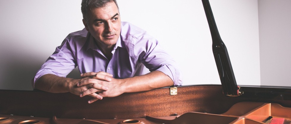 a man leans over the rim of an open grand piano, his hands clasped and the sleeves of his lavender button-down rolled up