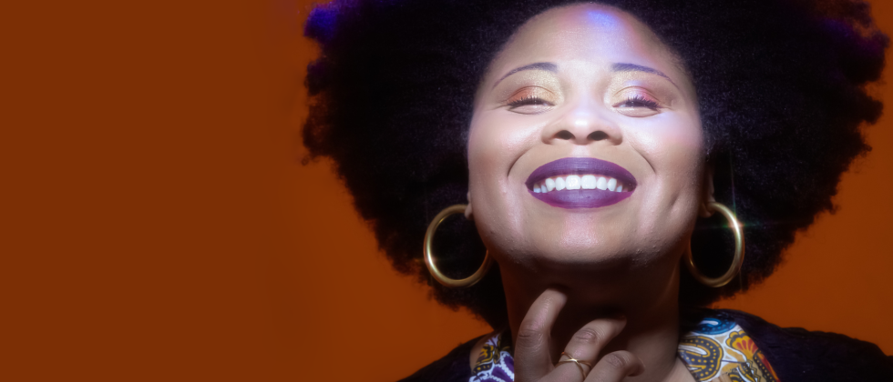 A woman with an afro smiles wearing large gold hoop earrings, a brightly colored scarf, and purple lipstick. 