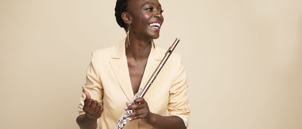 A black woman in a light tan jacket looks to the side, holding her flute in one hand across her body. She smiles warmly.