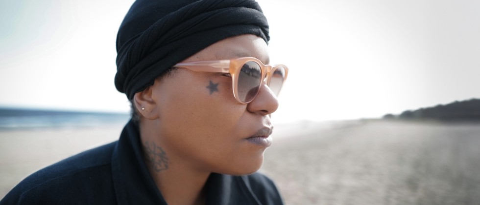 A black woman with a black knit cap, black collared shirt, and sunglasses looks into the distance outdoors. 