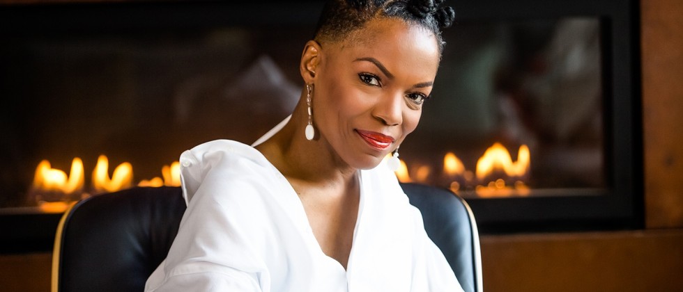 Nneena Freelon, a black woman in a white top, sits in front of a fire place and smiles.