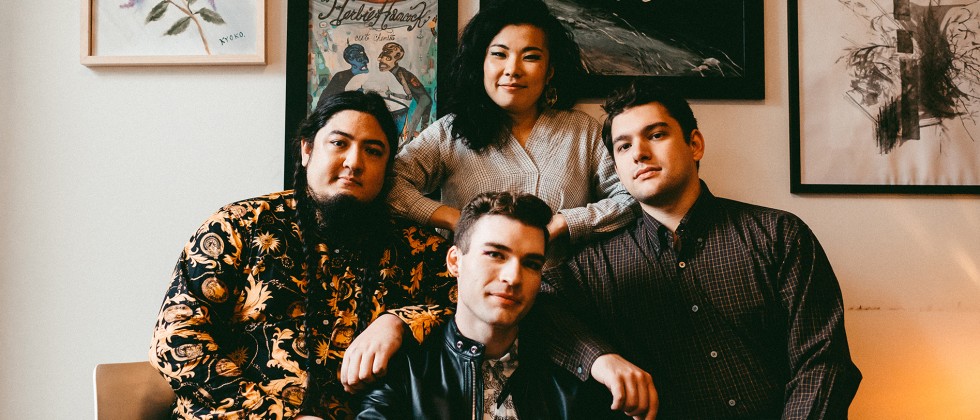 Four musicians stand close together, each member wears dark clothes with patterns, and three are holding a string instrument. 