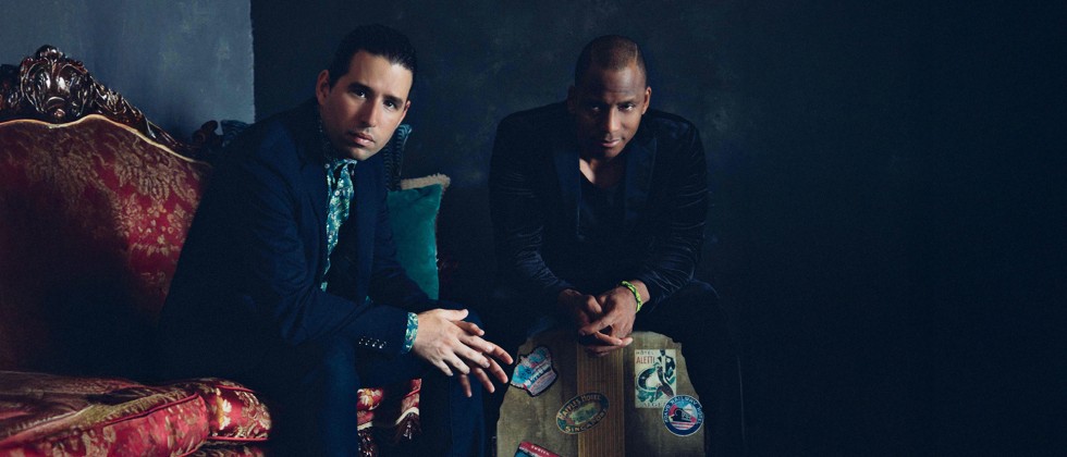 Two Cuban musicians sit in a dark room staring straight ahead, one is on an ornate red couch, one is on a vintage travel suitcase. 