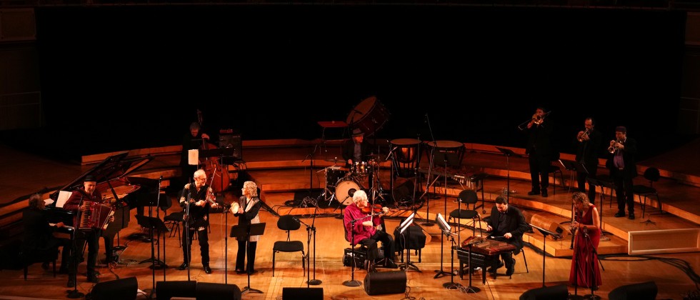 A large group of musicians are spread across a tiered stage with an older man in a red shirt plays the violin on a scooter power chair.