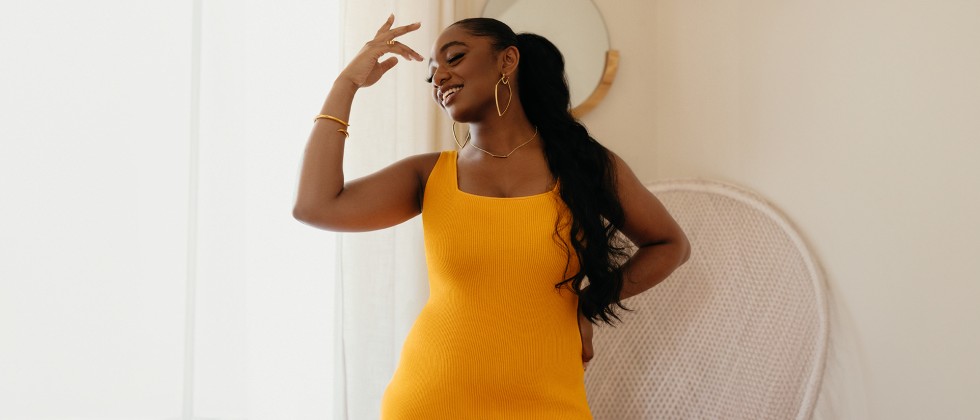 A young, Black woman wears a yellow, form fitting dress with gold jewelry. 