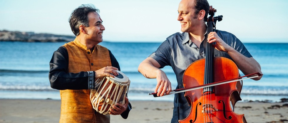 a south asian man holding one small drum and a white man bowing a cello stand on the beach, smiling at each other