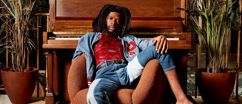 A Black man lounges shoeless in a textured armchair in front of an upright piano, surrounded by plants. 
