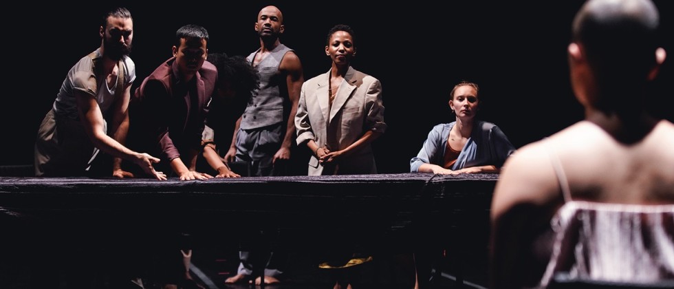 a diverse group of five men and women lean over a long table, as if challenging a person whose back is turned to us